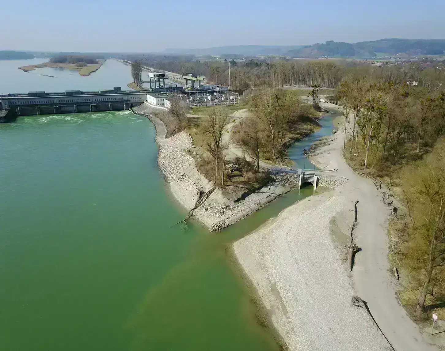 Luftaufnahme eines Kraftwerks an einem Fluss mit aufgestautem Wasser, angrenzendem Waldgebiet und Wanderwegen bei klarem Wetter.