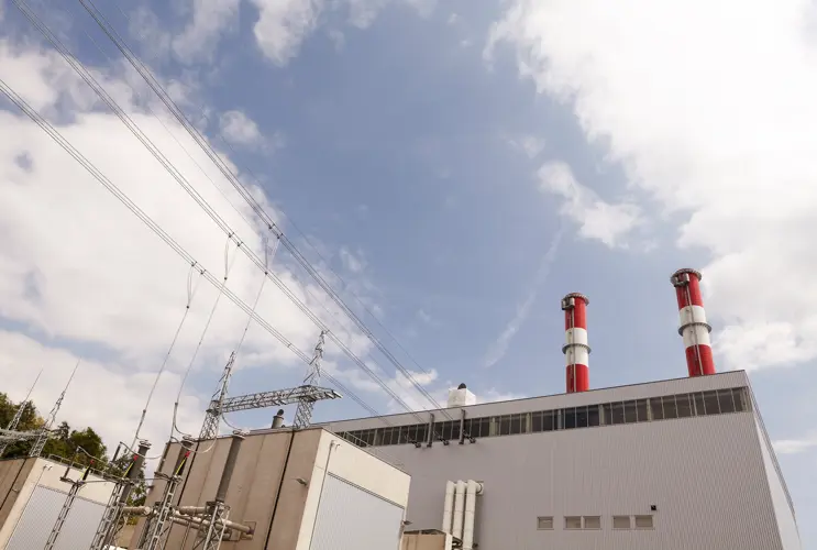 Wir blicken von unten auf das Gebäude des Wärmekraftwerks Mellach. Wir sehen Stromleitungen und zwei Schornsteine. Der Himmel ist blau.