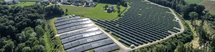 Ein Blick von oben auf eine großflächige Photovotaikanlage. Der Solarpark ist von sattem grün umgeben. Im Hintergrund sieht man kleinere malerische Ortschaften.