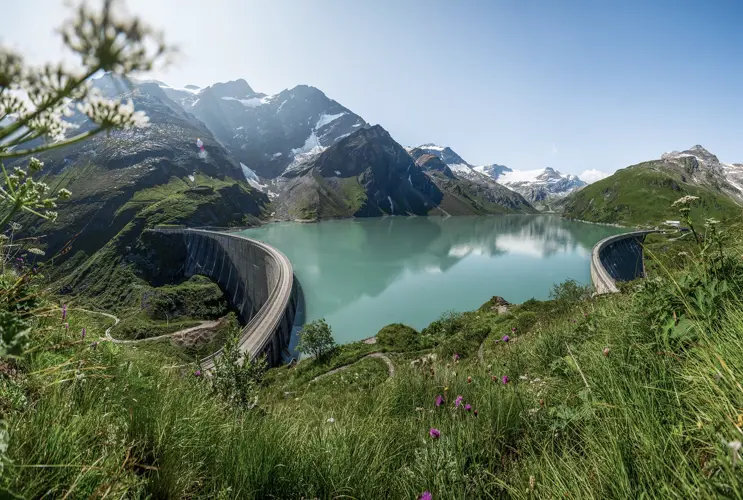 Ein Blick auf den Staussee bei Kaprun. Im Vordergrund sind Wiesen und Blumen zu sehen. Im Hintergrund schneebedeckte Berge.