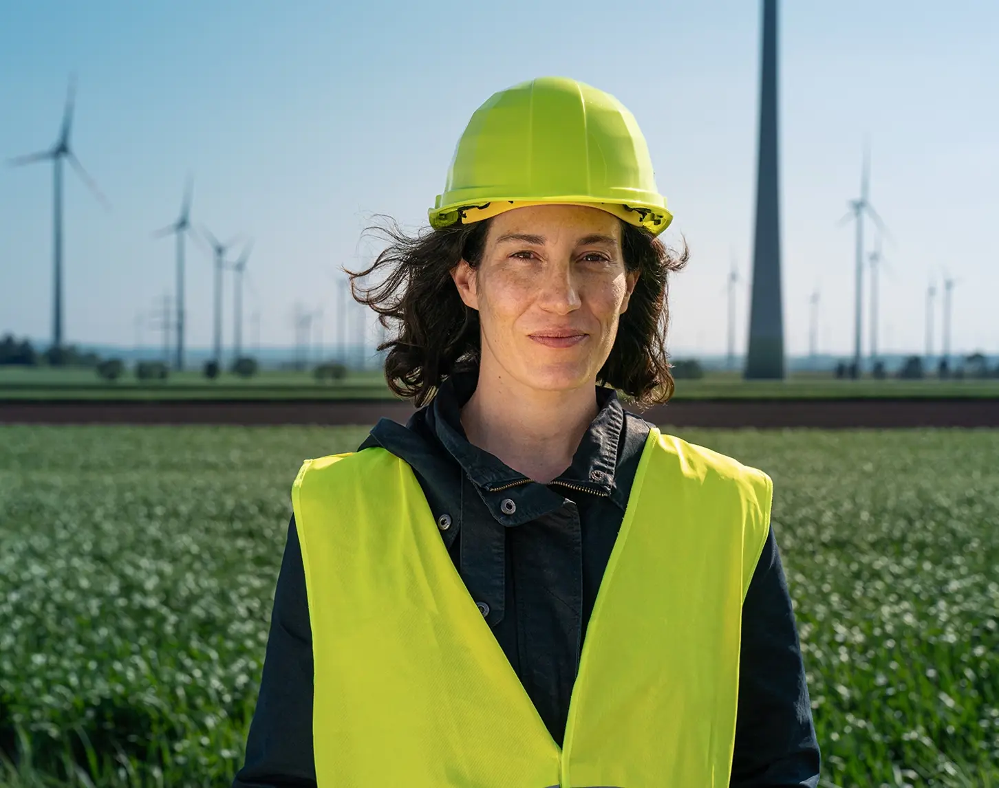 Eine Mitarbeiterin aus dem Bereich Windkraft steht in Sicherheitskleidung vor einem Windrad. Sie lächelt in die Kamera.