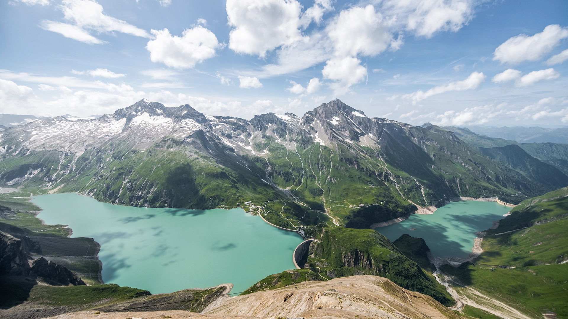 Panoramaansicht der Stauseen in Kaprun, umgeben von majestätischen Bergen und üppigem Grün. Die türkisfarbenen Seen liegen eingebettet in die alpine Landschaft und illustrieren die Besuchszentren von VERBUND.