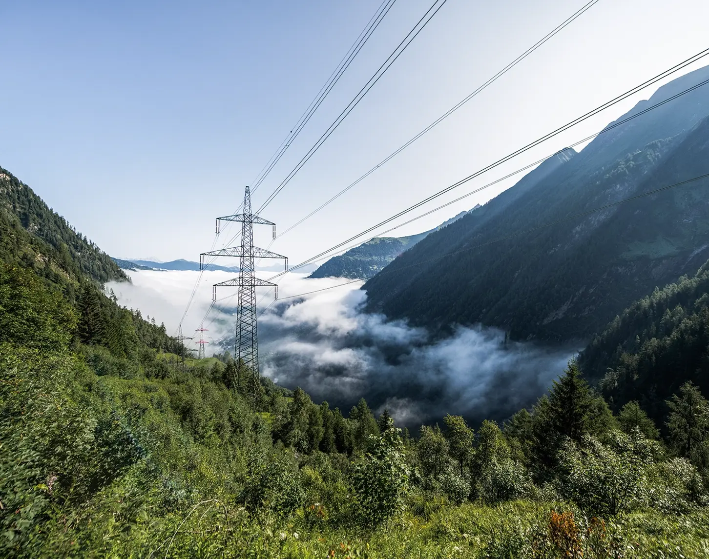 Zwischen teilweise bewalteten Hügeln steigen Nebenschwaden auf. Dazwischen sieht man ein Hochspannungsnetz.